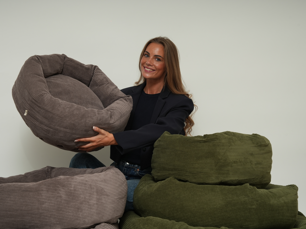 Woman holding a taupe corduroy dog bed beside a stack of moss green beds