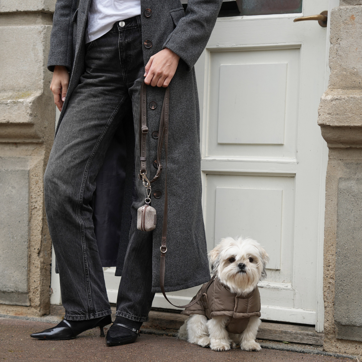 Een shih tzu aan de riem in een taupekleurige gewatteerde jas naast zijn baasje