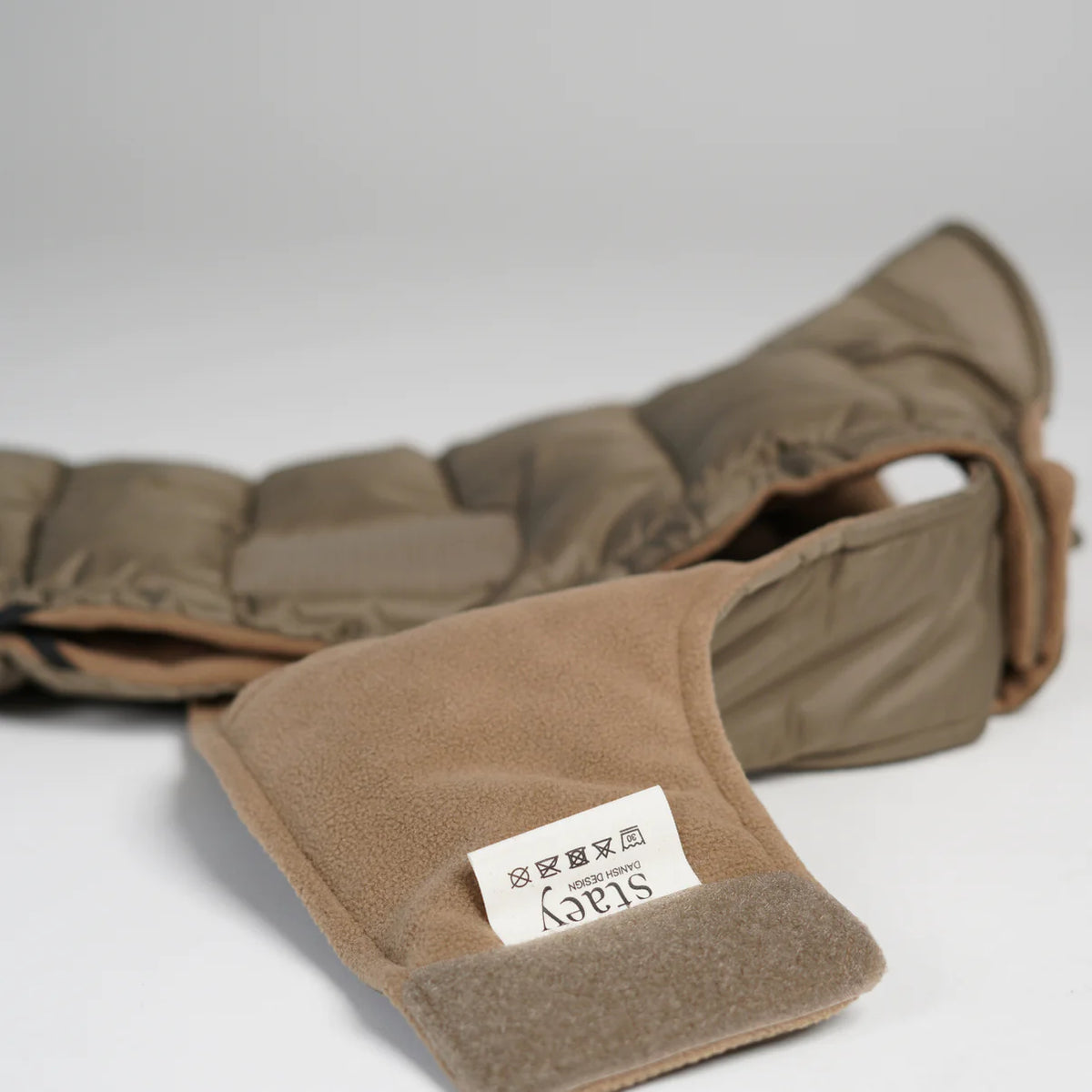 De taupekleurige, gewatteerde hondenjas ligt plat neer, waardoor de fleecevoering en het label zichtbaar zijn